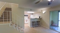 This two-story residence offers a well-organized layout that balances functionality and comfort across both levels. The main living area is finished with plush carpeting and flows naturally into a kitchen defined by light wood cabinetry, white appliances including a range, microwave, and refrigerator, and easy-care tile flooring. A peninsula-style breakfast bar with a granite-look countertop separates the kitchen from the living space, providing a casual seating area with room for bar stools — ideal for dining or conversation. Overhead, a textured ceiling adds subtle dimension throughout the main level, while wall-mounted lighting contributes a warm ambient glow to the space. A staircase with a white railing connects the main floor to the upper level, suggesting additional living or sleeping quarters above. HVAC vents are integrated discreetly into the ceiling, ensuring year-round climate comfort. The neutral color palette of soft greens and warm beige tones throughout creates a calm, welcoming atmosphere. Overall, the space is thoughtfully designed to maximize usability while maintaining an inviting, cohesive aesthetic across both floors.