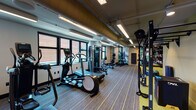This modern fitness center is thoughtfully designed to support a full range of workout routines within a single, open-plan floor. A row of large windows lines one wall, allowing ample natural light to fill the space and offering views of the surrounding brick exterior streetscape, creating an energizing atmosphere throughout.

The cardio zone is well-stocked with multiple treadmills, elliptical machines, rowing machines, and a stair climber, all positioned along the windowed wall to take advantage of the natural light. Nearby, a dedicated strength training area features a Precor cable machine, a multi-functional weight bench, and additional resistance equipment suited for targeted muscle training.

A separate functional training zone is distinguished by vibrant yellow flooring and bold, large-scale abstract wall murals in green and black tones. This area houses a comprehensive set of kettlebells, medicine balls, dumbbells arranged on tiered racks, and a Torque functional trainer, supporting dynamic and high-intensity workouts. A large flat-screen display and wall-mounted mirror enhance this zone further.

The ceiling features exposed ductwork and track lighting alongside flush linear fixtures, contributing to an industrial-modern aesthetic. Natural wood wall panels add warmth, while striped carpet flooring in the main cardio and strength areas provides a clean, professional finish throughout the space.