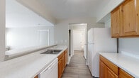 This single-floor unit offers a practical and comfortable layout across a well-utilized footprint. The galley-style kitchen is a functional centerpiece, featuring warm honey-toned wood cabinetry on both sides, light speckled countertops, white appliances including a refrigerator, electric range, and dishwasher, and a stainless steel sink. Wood-look flooring runs throughout the kitchen and hallway, adding a clean, durable contrast to the softer areas of the unit.

A pass-through opening connects the kitchen to the living area, which is carpeted in a neutral beige tone and receives ample natural light through a sliding glass door and adjacent window overlooking greenery. Vertical blinds provide privacy and light control in this space.

The bedroom is a well-proportioned room with plush carpet, a window with views of surrounding trees, and neutral walls that contribute to a calm atmosphere. A dedicated hallway connects the main areas, with two bathrooms accessible along its path, offering added convenience for guests and residents alike. Throughout the unit, a consistent neutral color palette and clean white trim create a cohesive, move-in-ready aesthetic.