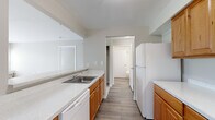 This single-floor unit offers a practical and comfortable layout across a well-utilized footprint. The galley-style kitchen is a functional centerpiece, featuring warm honey-toned wood cabinetry on both sides, light speckled countertops, white appliances including a refrigerator, electric range, and dishwasher, and a stainless steel sink. Wood-look flooring runs throughout the kitchen and hallway, adding a clean, durable contrast to the softer areas of the unit.

A pass-through opening connects the kitchen to the living area, which is carpeted in a neutral beige tone and receives ample natural light through a sliding glass door and adjacent window overlooking greenery. Vertical blinds provide privacy and light control in this space.

The bedroom is a well-proportioned room with plush carpet, a window with views of surrounding trees, and neutral walls that contribute to a calm atmosphere. A dedicated hallway connects the main areas, with two bathrooms accessible along its path, offering added convenience for guests and residents alike. Throughout the unit, a consistent neutral color palette and clean white trim create a cohesive, move-in-ready aesthetic.