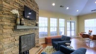 This inviting single-floor space exudes a warm and comfortable ambiance, thoughtfully designed for relaxation and social interaction. The focal point of the room is a dramatic floor-to-ceiling stacked stone fireplace, complete with a classic mantle and a wall-mounted flat-screen television above — a perfect blend of rustic charm and modern convenience. Vaulted ceilings enhance the sense of openness, adding an airy quality to the room. Natural light floods the interior through a series of large double-hung windows dressed with white horizontal blinds, offering views of the lush greenery outside. A glass-paneled exterior door further connects the interior to the outdoors. The space is furnished with plush navy blue armchairs and a coordinating sofa, arranged around a patterned area rug in earthy green and cream tones. A burnt orange upholstered ottoman adds a pop of color and serves as an additional seating or surface option. Warm hardwood flooring runs throughout, complementing the golden yellow walls and contributing to the overall cozy atmosphere. Recessed lighting provides a soft, even glow, making this a versatile and welcoming gathering space at any time of day.