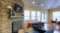 This inviting single-floor space exudes a warm and comfortable ambiance, thoughtfully designed for relaxation and social interaction. The focal point of the room is a dramatic floor-to-ceiling stacked stone fireplace, complete with a classic mantle and a wall-mounted flat-screen television above — a perfect blend of rustic charm and modern convenience. Vaulted ceilings enhance the sense of openness, adding an airy quality to the room. Natural light floods the interior through a series of large double-hung windows dressed with white horizontal blinds, offering views of the lush greenery outside. A glass-paneled exterior door further connects the interior to the outdoors. The space is furnished with plush navy blue armchairs and a coordinating sofa, arranged around a patterned area rug in earthy green and cream tones. A burnt orange upholstered ottoman adds a pop of color and serves as an additional seating or surface option. Warm hardwood flooring runs throughout, complementing the golden yellow walls and contributing to the overall cozy atmosphere. Recessed lighting provides a soft, even glow, making this a versatile and welcoming gathering space at any time of day.