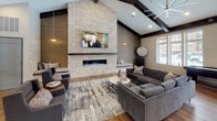 This single-floor community room is a well-appointed shared gathering space that seamlessly blends modern design with warm, inviting elements. The architecture is defined by dramatic vaulted ceilings accented with dark exposed wood beams, creating a sense of openness and volume throughout the entire space.

The fully equipped kitchen serves as a functional centerpiece, featuring dark cabinetry with a herringbone-pattern tile backsplash, stainless steel appliances including a gas range, double ovens, a refrigerator, and a microwave. A large white-topped island with bar seating provides an ideal spot for casual dining or socializing, complemented by globe pendant lighting overhead. An adjacent high-top dining table with bar-height chairs offers additional seating alongside a wall-mounted television.

The lounge area is anchored by a stunning floor-to-ceiling stacked stone accent wall with an integrated linear fireplace and a mounted flat-screen television, creating a dramatic focal point. Plush gray sectional sofas are arranged around a patterned area rug, fostering a relaxed and comfortable atmosphere. A large ceiling fan adds to the room's comfort, while an expansive bank of windows along one wall floods the space with natural light and offers views of the surrounding neighborhood. Warm hardwood flooring runs throughout, tying the kitchen, dining, and lounge areas together cohesively.