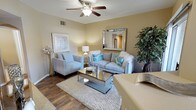 This single-floor space showcases a thoughtfully designed living area that balances comfort and style. The living room serves as the focal point, finished with rich hardwood-style flooring that grounds the space and contrasts elegantly with the warm, neutral-toned walls painted in a soft beige. A ceiling fan with an integrated light fixture overhead provides both practical airflow and ambient lighting throughout the room.

The seating arrangement features a plush sofa and a coordinating armchair, both upholstered in light silver-gray fabric and accented with teal and white decorative pillows that introduce a pop of color. A glass and metallic coffee table sits atop a layered area rug, adding a contemporary, reflective element to the composition. A large framed mirror mounted on the wall enhances the sense of space and light, while a tall decorative indoor tree adds a natural, organic touch to the corner. Subtle artwork and decorative accessories complete the styling. The overall ambiance is polished yet welcoming, with a cohesive color palette that feels modern and livable.