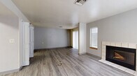 This single-floor unit offers an efficient and functional layout thoughtfully designed to maximize its compact footprint. The combined kitchen and living room serves as the heart of the space, centered around warm wood-look laminate flooring that extends throughout. The living area features a tile-surround fireplace as a focal point, flanked by a window with blinds and a sliding glass door fitted with vertical blinds that invites natural light and provides access to the outdoors. The kitchen is well-appointed with warm-toned wood cabinetry, granite-style countertops, and a full suite of stainless steel appliances including a range, microwave, dishwasher, and refrigerator, all illuminated by overhead fluorescent lighting. Three bathrooms serve the unit, each finished with light tile flooring. One bathroom features a generous vanity with a granite countertop, an undermount sink, and wood cabinetry, complemented by a large framed mirror. Additional bathrooms offer clean, utilitarian designs with white tile flooring. The neutral gray and white color palette throughout contributes to a clean, modern aesthetic, while multiple ceiling vents ensure consistent climate control. The open dollhouse view reveals a well-organized floor plan that balances living, cooking, and bathroom spaces efficiently within the available area.