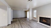 This single-floor unit offers an efficient and functional layout thoughtfully designed to maximize its compact footprint. The combined kitchen and living room serves as the heart of the space, centered around warm wood-look laminate flooring that extends throughout. The living area features a tile-surround fireplace as a focal point, flanked by a window with blinds and a sliding glass door fitted with vertical blinds that invites natural light and provides access to the outdoors. The kitchen is well-appointed with warm-toned wood cabinetry, granite-style countertops, and a full suite of stainless steel appliances including a range, microwave, dishwasher, and refrigerator, all illuminated by overhead fluorescent lighting. Three bathrooms serve the unit, each finished with light tile flooring. One bathroom features a generous vanity with a granite countertop, an undermount sink, and wood cabinetry, complemented by a large framed mirror. Additional bathrooms offer clean, utilitarian designs with white tile flooring. The neutral gray and white color palette throughout contributes to a clean, modern aesthetic, while multiple ceiling vents ensure consistent climate control. The open dollhouse view reveals a well-organized floor plan that balances living, cooking, and bathroom spaces efficiently within the available area.