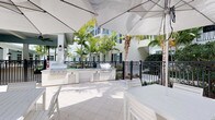 This inviting outdoor communal space is designed for relaxed, resort-style living. At the heart of the area is a well-appointed outdoor kitchen, complete with multiple stainless steel grills, ample counter space, and built-in cabinetry — everything needed for effortless outdoor entertaining. Large sail shade canopies stretch overhead, providing generous sun coverage over the tiled patio and its crisp white outdoor furnishings, creating a cool and comfortable gathering space even on the sunniest days. Tall, elegant palm trees frame the area, enhancing the tropical ambiance and adding a lush, verdant backdrop. The patio is enclosed by classic wrought iron fencing, lending a refined aesthetic that complements the architectural style of the surrounding multi-story buildings. Lantern-style wall sconces and a ceiling fan on the adjacent covered walkway add both charm and function. The overall atmosphere is bright, clean, and welcoming, with a cohesive design that blends coastal influences with upscale communal amenities, making this an ideal spot for gatherings, cookouts, or simply enjoying the warm, tropical setting.