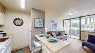 This single-floor, high-rise unit offers a practical and cohesive layout designed to maximize comfort and functionality. The kitchen is equipped with white cabinetry, a white electric range, a refrigerator, and a sink, all set against wood-look flooring. A peninsula breakfast bar with seating extends into the living and dining area, creating a seamless open-concept feel that encourages easy interaction between spaces.

The living area is carpeted and features a distinctive white tile-panel accent wall that adds texture and visual interest to the room. Large windows dressed with vertical blinds allow ample natural light to filter in, with views of surrounding greenery. The space is well-suited for both relaxing and entertaining.

The bedroom is generously sized, also carpeted, and benefits from an oversized window that fills the room with daylight. The white tile-panel wall continues here, maintaining design consistency throughout the unit.

The bathroom features wood-look flooring, a vanity with under-sink cabinetry, tiled walls, and a mirror above the sink, providing a clean and functional space. Overall, the unit presents a bright, neutral palette with consistent finishes that create a cohesive and livable environment.