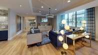 This thoughtfully designed lobby offers a welcoming and sophisticated environment across a single open floor. The space is anchored by a generously sized lounge area furnished with a striking royal blue sectional sofa, complementary patterned accent chairs, and layered area rugs in coordinating blue and white tones. An elegant crystal chandelier and stylish globe floor lamps provide warm, layered lighting throughout the seating area, while a wall-mounted flat-screen display adds a modern, functional touch above a decorative credenza.

Adjacent to the lounge, a dining or collaboration zone features a rich wood high-top table surrounded by upholstered bar-height chairs, ideal for casual meetings or social gatherings. Large floor-to-ceiling windows dressed with patterned drapery flood both areas with natural light and offer views of the surrounding streetscape.

A dedicated beverage station serves as a practical amenity, outfitted with navy blue cabinetry, granite countertops, pendant lighting, a textured subway tile backsplash, and a coffee machine — providing a refined and convenient refreshment area for residents or guests.

Light hardwood flooring runs throughout the entire space, tying together the open-plan layout. Decorative sculpted elements, artistic wall features, and ornate metalwork railings further elevate the lobby's contemporary aesthetic, creating a polished and inviting first impression.