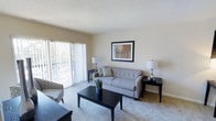 This single-floor residential space offers a comfortable and well-appointed layout designed for everyday living. The living room serves as the heart of the home, featuring plush wall-to-wall carpeting that adds warmth underfoot and a soft, neutral color palette that creates a calm, inviting atmosphere. A textured ceiling adds subtle visual interest overhead.

Natural light flows generously into the living area through large sliding glass doors dressed with vertical blinds, which also provide access to an outdoor space and offer views of lush greenery beyond. The room comfortably accommodates a full seating arrangement, including a tufted sofa, accent chair, and a dark-finished coffee table, all thoughtfully arranged to foster an open, functional flow.

Warm table lamps on either side of the seating area contribute to a cozy ambiance during the evening hours, while framed wall art adds a finishing decorative touch. The overall design of the space strikes a balance between simplicity and comfort, making it an ideal setting for both relaxing and entertaining. The light tones of the walls and flooring work together to enhance the sense of openness throughout the home.