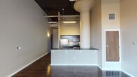 This single-floor urban loft showcases an industrial-chic aesthetic defined by dramatic ceiling height, exposed ductwork, and structural columns that add architectural character throughout the space. Rich, dark hardwood floors run continuously across the open-concept living and kitchen areas, grounding the design with warmth and depth.

The kitchen is a standout feature, equipped with stainless steel appliances including a gas range, over-the-range microwave, and refrigerator. Dark espresso cabinetry pairs elegantly with granite countertops, while a mosaic tile backsplash adds a refined decorative detail. A peninsula with additional counter space and a double sink provides both functionality and a natural visual divide between the kitchen and living areas.

The living area is anchored by a striking arched window that floods the space with natural light and offers views to the exterior. The open ceiling with its dark-painted industrial finish amplifies the sense of volume and loftiness in this area.

The bedroom offers a quieter retreat, maintaining the same dark hardwood flooring and neutral wall tones, with the industrial ceiling details continuing throughout. The bathroom is well-appointed with dark vanity cabinetry, granite countertops, a large mirror with vanity lighting, and a tiled tub and shower surround, rounding out this thoughtfully designed urban residence.