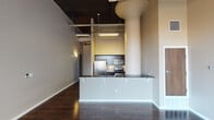 This single-floor urban loft showcases an industrial-chic aesthetic defined by dramatic ceiling height, exposed ductwork, and structural columns that add architectural character throughout the space. Rich, dark hardwood floors run continuously across the open-concept living and kitchen areas, grounding the design with warmth and depth.

The kitchen is a standout feature, equipped with stainless steel appliances including a gas range, over-the-range microwave, and refrigerator. Dark espresso cabinetry pairs elegantly with granite countertops, while a mosaic tile backsplash adds a refined decorative detail. A peninsula with additional counter space and a double sink provides both functionality and a natural visual divide between the kitchen and living areas.

The living area is anchored by a striking arched window that floods the space with natural light and offers views to the exterior. The open ceiling with its dark-painted industrial finish amplifies the sense of volume and loftiness in this area.

The bedroom offers a quieter retreat, maintaining the same dark hardwood flooring and neutral wall tones, with the industrial ceiling details continuing throughout. The bathroom is well-appointed with dark vanity cabinetry, granite countertops, a large mirror with vanity lighting, and a tiled tub and shower surround, rounding out this thoughtfully designed urban residence.