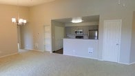 This single-floor residence offers a clean, functional layout designed to maximize comfort and usability. The living area is finished with plush wall-to-wall carpeting, creating a warm and inviting atmosphere. A built-in shelving nook is recessed into the wall, providing practical display or storage space without sacrificing floor area. The living space flows seamlessly into the kitchen through an open pass-through design, enhancing the sense of connectivity between rooms. The kitchen is equipped with stainless steel appliances, including a refrigerator, range with oven, and an over-the-range microwave. Gray cabinetry lines the upper walls, offering generous storage, while light-toned countertops add a bright, contemporary contrast. Dark hardwood-style flooring in the kitchen provides a clean visual distinction from the carpeted living space. A pantry or closet with a paneled door is accessible from the kitchen area, adding to the overall storage capacity. The neutral color palette throughout — soft beige walls, light countertops, and gray cabinetry — creates a cohesive and versatile backdrop suited to a variety of interior styles. Overall, the space is well-organized, efficiently designed, and move-in ready.
