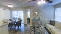 This single-story space presents a thoughtfully designed interior that balances comfort and style. The main living and dining area is anchored by rich, dark hardwood flooring that extends throughout, complemented by neutral beige and soft blue-gray walls that create a calm, welcoming atmosphere. A ceiling fan with a light fixture overhead provides both function and a subtle decorative touch.

The dining area accommodates a dark wood table with upholstered chairs, positioned near large sliding glass doors that invite abundant natural light and offer views of the surrounding outdoor greenery. This connection to the outside enhances the open, airy feel of the space.

A decorative starburst mirror with metallic detailing serves as a striking focal point on the accent wall, flanked by teal decorative accessories atop a silver mirrored console. A glass-topped coffee table and a patterned area rug add layers of texture to the living area.

A doorway visible from the main room leads to an adjacent bedroom or private space, also benefiting from natural light through its own window. Chair rail molding along the walls adds an architectural detail that elevates the overall design aesthetic.