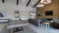 This single-floor amenity space is designed as a social and recreational lounge, combining contemporary style with inviting comfort. The room immediately draws attention with its soaring vaulted ceiling, accented by dark exposed wooden beams that add architectural character and a rustic-modern contrast against the crisp white walls. Recessed lighting is complemented by striking oversized circular pendant fixtures, providing warm, layered illumination throughout the space.

A full-size billiards table serves as the room's centerpiece, positioned on a neutral-toned area rug and surrounded by ample space for gameplay. Adjacent to the table, a comfortable gray sectional sofa offers relaxed seating, arranged around a wooden side table with decorative greenery and accessories. A wall-mounted flat-screen television is positioned above this lounge area, making it well-suited for both casual gatherings and entertainment.

A standout feature is the full-height wood-paneled accent wall, which introduces natural warmth and texture to the space. A sliding barn door leads to an adjacent room, adding both a functional and stylish architectural detail. The overall ambiance is polished yet approachable, ideal for communal recreation and socializing.