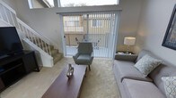This two-story residence offers a well-organized layout designed for comfortable everyday living. The living room serves as the heart of the main floor, featuring plush carpeting and an abundance of natural light streaming in through large sliding glass doors fitted with vertical blinds. These doors open directly onto an outdoor patio space, visible through the glass and surrounded by neighboring structures and mature palm trees, providing a pleasant connection to the outdoors. The living area is generously proportioned, accommodating a sofa, accent chair, and a central coffee table with ease. A staircase positioned along one wall leads to the upper floor, featuring carpeted treads and a clean white railing. High transom windows above the sliding doors draw in additional light, enhancing the open and airy feel of the main living space. A side table with a lamp adds a warm, functional touch to the room. The overall aesthetic is clean and neutral, with white walls and light-toned finishes throughout. The second floor provides additional living space, accessible via the interior staircase, making this a practical and versatile multi-level home.