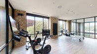 This dedicated cardio fitness room offers a thoughtfully designed workout environment that balances industrial charm with modern aesthetics. Exposed brick columns punctuate the space, contrasting beautifully with smooth white ceilings fitted with recessed and in-ceiling speakers. Light hardwood-style flooring runs throughout, lending warmth and continuity to the room.

Floor-to-ceiling windows and sliding glass doors line the perimeter, flooding the space with abundant natural light and offering open views of the surrounding greenery and neighboring buildings. A large glass partition wall with dark steel framing separates sections of the room while maintaining visual openness.

The room is outfitted with a curated selection of cardiovascular and strength equipment, including multiple stationary cycling bikes, rowing machines, and a well-stocked kettlebell rack in a range of weights. A wall-mounted mat and a dumbbell storage rack provide additional functional fitness options. A large flat-screen television is built into a warm wood-panel accent wall, ideal for guided workouts or entertainment during training sessions.

A full-length mirror further enhances the sense of space and supports proper form during exercise. The overall ambiance is clean, bright, and professional, creating an inviting atmosphere for focused and effective cardio training.