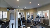 This fitness center is designed to support a well-rounded workout routine, offering a thoughtful mix of strength training and cardiovascular equipment within a single-floor layout. The space features a Life Fitness multi-station strength machine prominently positioned near one wall, accompanied by an exercise reference chart for guided use. Cardio equipment includes a stationary upright bike, a treadmill, and an elliptical trainer, providing a range of aerobic workout options. The flooring is finished in a dark rubberized material, practical and durable for high-traffic fitness use. Recessed lighting is evenly distributed across the smooth, light-toned ceiling, ensuring the space is well-lit throughout. Natural light is introduced through windows along one wall, adding brightness and openness to the environment. Glass-paneled interior doors provide a visual connection to an adjacent room, enhancing the sense of depth. The walls are painted in a neutral beige tone, contributing to a clean, functional atmosphere. A posted notice board is visible near the entrance, indicating shared-use guidelines for the space. Overall, the fitness center maintains a practical and organized environment suited for everyday use.