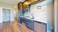 Located at 120 West Ave in Kannapolis, NC, this modern unit offers a well-designed single-floor layout comprising a kitchen, bathroom, hallway, and entrance. The open living and kitchen area is anchored by warm wood-look flooring, neutral gray walls, and a sleek ceiling fan that enhances the airy, contemporary feel of the space. A full-width sliding glass door floods the interior with natural light and opens onto a private balcony offering open views of the surrounding Kannapolis cityscape and greenery. The kitchen is a standout feature, outfitted with dark wood-grain cabinetry extending to the ceiling for ample storage, stainless steel appliances including a refrigerator, range, built-in microwave, and dishwasher, along with clean white countertops and an eye-catching herringbone marble tile backsplash. A matte black pull-down faucet adds a refined touch. The bathroom is finished with large-format gray floor tiles, a glass-enclosed walk-in shower with matching tile surround, matte black hardware and fixtures, and an in-unit stacked washer/dryer. The entrance and hallway connect the spaces efficiently, contributing to a cohesive, functional flow throughout the unit.