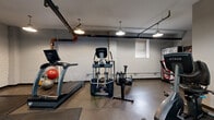 This exercise room offers a fully functional fitness environment within a compact, efficiently utilized footprint. The space features an industrial aesthetic, defined by exposed overhead pipes—including distinctive orange-accented plumbing—dark ceiling ducts, and pendant-style dome lights that cast warm, focused illumination throughout. The walls are finished in painted concrete block, contributing to the room's utilitarian character, while a small window provides a modest source of natural light.

The equipment layout is thoughtfully organized to maximize usable floor space. Cardio offerings include a True-brand treadmill, an elliptical trainer, a rowing machine, and a stationary bike. Strength training is well-represented with a power rack loaded with an Olympic barbell and weight plates, a full dumbbell rack, and an assortment of exercise balls for functional fitness and stretching. Rubber floor mats are positioned beneath equipment for both safety and floor protection.

A large wall mirror enhances the sense of space and allows for form-checking during workouts. Glass-paneled doors provide access to an adjacent area, adding openness to the layout. Overall, the room delivers a practical, no-frills fitness experience with a solid range of equipment suited for both cardiovascular and strength-based training routines.