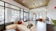 Located at 120 Whitby Lane in Birmingham, Alabama, this thoughtfully designed amenity space seamlessly blends sophisticated interior living with an expansive outdoor balcony experience. The interior lounge area is anchored by warm wood-plank ceilings with recessed lighting, light hardwood flooring, and crisp white shiplap and textured accent walls. A stylish kitchenette runs along one side, featuring sleek gray quartz countertops, dark cabinetry, a black matte sink, and pendant lighting overhead — ideal for entertaining. The lounge itself is outfitted with a generous sectional sofa, a mounted flat-screen television, and an open layout that encourages comfortable gathering. Large steel-framed glass doors connect the interior to the covered balcony, flooding the space with abundant natural light. The balcony extends the living area outdoors, where tall latticed windows frame views of the surrounding community, lush greenery, and the Birmingham skyline in the distance. A substantial dining table with woven chairs sits beneath a ceiling fan, making it a functional and inviting al fresco setting. Manicured planters line the balcony perimeter, adding a layer of privacy and natural beauty. The cohesive design palette of white, gray, and natural wood tones throughout creates a refined yet welcoming atmosphere.