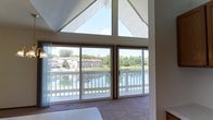 This single-floor space makes a striking architectural statement with its vaulted ceiling that rises to a sharp peak, framing a large triangular clerestory window at the apex. The design maximizes natural light, flooding the interior with brightness throughout the day. Beneath the vaulted ceiling, a wall of sliding glass doors spans the width of the main living area, providing unobstructed panoramic views of a serene pond or lake. The sliding doors open directly onto a balcony enclosed by white railings, creating a seamless transition between the interior and the outdoor setting. Lush greenery and mature trees line the far bank of the water, while neighboring buildings are visible in the background, adding a sense of community to the surroundings. The interior features neutral-toned carpet underfoot, complementing the warm wood-framed windows and the soft, earthy color palette of the walls. The combination of the dramatic vaulted ceiling, abundant natural light, and captivating waterfront views gives the space an open, airy ambiance that is both inviting and visually impressive.
