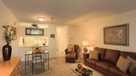This single-floor residence offers a welcoming open-concept layout that seamlessly connects the main living area to the kitchen. The living room is carpeted and comfortably sized, accommodating a rich brown upholstered sofa and matching armchair with ottoman, centered around a glass-topped coffee table. Warm ambient lighting from a ceiling fixture and a table lamp creates an inviting atmosphere throughout the space.

A pass-through or breakfast bar connects the living area to the kitchen, which features light-colored cabinetry and countertops, with a window above the sink that brings in natural light. A compact dining area with a glass and iron table and chairs is positioned near the kitchen, making it practical for everyday meals.

Neutral, cream-colored walls complement the beige carpeting, contributing to the overall warm and understated aesthetic. A hallway visible from the main living area leads to additional rooms, hinting at a practical and well-organized floor plan. Framed artwork on the walls adds a finishing touch, enhancing the lived-in comfort of the space.