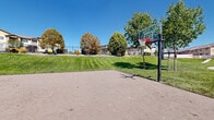 This community recreational space offers a thoughtfully designed outdoor environment suited for active outdoor living. At its center is a concrete basketball court equipped with a sturdy regulation-height hoop featuring a clear backboard, providing an ideal surface for recreational play. Adjacent to the court, a sand court area offers additional recreational possibilities, while expansive, well-manicured green lawns stretch across the grounds, creating open space for leisure and play.

Winding concrete pathways meander through the landscaped terrain, connecting the various activity areas and inviting residents to walk or jog through the grounds. The gently rolling landscape adds visual interest and natural separation between zones. Mature deciduous trees are scattered throughout, providing shade and seasonal color, while ornamental plantings and rock features contribute to the overall aesthetic appeal.

The surrounding residential buildings frame the space, giving it a neighborhood park character. Panoramic views of distant mountain ranges can be seen on clear days, adding a scenic backdrop to outdoor activities. The entire space benefits from abundant natural light under open skies, making it an inviting destination year-round for recreation and relaxation.