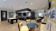This single-floor recreation and social lounge is thoughtfully designed to accommodate a wide range of leisure activities in a cohesive, stylish environment. The centerpiece of the space is a striking built-in bar area, clad in dark navy shiplap paneling with a rich wood-toned countertop and anchored by a coffered ceiling detail overhead. A row of industrial-style barstools lines the bar, which also features a wall-mounted television — ideal for gathering around during events or game days. Adjacent to the bar, open lounge areas are furnished with patterned yellow and neutral upholstered seating, round accent tables, and warm decorative lighting, creating an inviting social atmosphere. The game room section is expansive, housing a billiards table, an air hockey table, a ping-pong table, and a foosball table, all set against a backdrop of dark wainscoting and large windows that allow generous natural light to fill the space. A wall-mounted dartboard adds another recreational option. A dedicated lounge nook features bold yellow accent walls, wall-mounted televisions, framed black-and-white artwork, and cozy seating. Throughout, neutral flooring ties the zones together, while recessed lighting ensures the entire space is well-lit and welcoming.