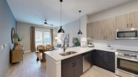 Located at 1100 Warburton Rd in Summerville, SC, this well-appointed 2-bedroom, 1-bathroom residence offers a thoughtfully designed single-floor layout. The open-concept kitchen is a standout feature, boasting a large quartz peninsula with bar seating, bold dark lower cabinetry contrasted by light wood upper cabinets, a white subway tile backsplash, and a full suite of stainless steel appliances including a refrigerator, range, microwave, and dishwasher. Striking black pendant lights hang above the peninsula, adding a modern industrial touch. The kitchen flows seamlessly into the living room, which is bathed in natural light from large sliding glass doors that open to an outdoor balcony, complemented by a ceiling fan overhead. Wood-look plank flooring runs throughout the main living areas and hallway, creating a warm and cohesive aesthetic. The primary bedroom features a soothing accent wall, a ceiling fan, and generous natural light from large windows. The bathroom is clean and contemporary, with a dark-framed glass walk-in shower, a dark vanity with quartz countertop, and wood-look tile flooring. Built-in shelving near the entry and a dedicated closet provide ample storage throughout the home.