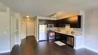 This single-floor residence offers a thoughtfully designed open-concept layout that seamlessly connects the kitchen and living areas. The kitchen is a standout feature, outfitted with sleek dark espresso cabinetry that provides ample storage and a bold visual contrast against the light-toned tile backsplash. Stainless steel appliances — including a refrigerator, range, microwave, and dishwasher — lend a modern, cohesive aesthetic. A double-basin sink with a contemporary faucet anchors the workspace, while track lighting overhead ensures the area is well-lit and functional. Rich, dark hardwood flooring extends throughout the space, adding warmth and a sense of continuity. The open flow from the kitchen into the adjacent hallway and connecting rooms makes the layout feel spacious and easy to navigate. Recessed and flush-mount ceiling fixtures provide consistent ambient lighting throughout. White paneled doors and clean white walls create a crisp, neutral backdrop that balances the darker finishes. The overall ambiance is modern and polished, combining practical design with stylish details to create a comfortable and visually cohesive living environment.