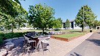 This thoughtfully designed outdoor amenity space offers residents a comprehensive recreational experience centered around a spacious paver patio. At the heart of the space is a built-in outdoor kitchen station constructed from stacked stone, topped with a dark granite countertop and equipped with a built-in grill. High-top bar seating lines the counter, providing a convenient spot for casual dining and socializing. Adjacent to the grilling area, a regulation-sized bocce ball court is framed by cedar wood borders and features a well-maintained artificial turf surface, offering a dedicated space for outdoor recreation. Comfortable Adirondack-style chairs are arranged nearby, creating a relaxed seating area ideal for conversation or unwinding. Further along the patio, additional picnic-style tables provide ample communal dining space. The entire area is shaded by mature deciduous trees, creating a cool and inviting atmosphere on sunny days. Lush landscaping, including ornamental shrubs and garden beds defined by natural stone edging, adds to the serene, park-like ambiance. The surrounding green lawn and open sky views complete this well-rounded outdoor retreat.