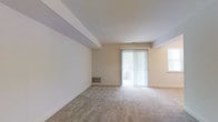 This single-floor residence offers a clean, functional layout with a calm and neutral aesthetic. The main living area is generously proportioned, featuring wall-to-wall carpeting in a warm beige tone that adds a soft, comfortable feel underfoot. The space is bathed in natural light, thanks to a large sliding glass door fitted with vertical blinds that opens directly onto a balcony, providing a seamless connection to the outdoors. An additional side window further enhances the brightness of the room. The ceiling features a subtle architectural beam detail that adds a touch of dimension to the otherwise smooth, light-toned surfaces. Walls are finished in a clean off-white, contributing to the airy and open ambiance. A floor-level HVAC vent is integrated into the baseboard, ensuring efficient climate control throughout the space. The overall design is straightforward and versatile, offering a blank canvas that adapts easily to a variety of furnishing styles. The combination of natural light, neutral tones, and practical features makes this a welcoming and livable space.