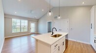 Located on Red Buckeye Drive in Huber Heights, Ohio, this single-floor, 2-bedroom, 2-bathroom home combines modern design with functional living. The open-concept kitchen and living area is a standout feature, boasting soaring vaulted ceilings, warm light wood-look flooring, and recessed lighting. The kitchen is well-appointed with crisp white shaker cabinetry, a white subway tile backsplash, a central island with a sink and pendant lighting, and black appliances. The living area receives generous natural light through large windows and offers direct outdoor access via a glass-paned door. Both bedrooms are finished with the same light wood flooring and neutral tones, with windows allowing natural light and outdoor views. The spacious primary bedroom includes a dedicated walk-in closet outfitted with wire shelving systems. The two bathrooms are finished with wood-look flooring, with one featuring a white vanity with a modern matte black faucet, a large mirror, and a bright window. The second bathroom includes a full tub/shower surround with built-in shelving and matte black fixtures. Consistent neutral wall tones and clean white trim unify the home's modern and welcoming aesthetic throughout.