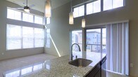 This single-floor space showcases a sleek, contemporary design with an open-concept layout that seamlessly connects the kitchen and living areas. The kitchen is a standout feature, highlighted by a generous granite countertop island with a stainless steel undermount sink and a gooseneck faucet. Pendant lighting in frosted cylindrical shades hangs gracefully above the island, adding both functional illumination and a modern aesthetic touch. Stainless steel appliances complement the refined finishes throughout.

One of the most striking aspects of the space is its impressive ceiling height, which draws the eye upward and contributes to an expansive, airy atmosphere. Large floor-to-ceiling windows and sliding glass doors flood the interior with abundant natural light, casting bright, sweeping rays across the living area. The windows are fitted with horizontal blinds for adjustable privacy and light control.

The living area features neutral-toned flooring and walls, providing a clean, versatile backdrop. A private balcony accessible through the glass doors offers outdoor views of the surrounding urban environment, including neighboring buildings. The overall ambiance is bright, modern, and open, making excellent use of natural light and architectural scale to create a welcoming and functional living space.