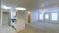 This single-floor space features an open-concept layout that seamlessly connects the kitchen and living area, creating a cohesive and functional environment. The kitchen is positioned to one side and includes white cabinetry, a dark countertop, and a pendant light fixture overhead that provides focused task lighting. A partial wall divides the kitchen from the broader living space without fully enclosing either area, maintaining an open feel throughout.

The living area is carpeted and benefits from natural light entering through a window on the far wall, contributing to a bright and welcoming atmosphere. A ceiling fan with a built-in light fixture is centrally located in the living space, offering both ventilation and ambient lighting. The walls throughout are finished in a neutral white, keeping the interior feeling clean and versatile.

Two interior doors are visible toward the back of the space, suggesting access to additional rooms or storage areas. The overall layout is straightforward and practical, with a neutral color palette and simple finishes that provide a blank canvas adaptable to a variety of furnishing styles and preferences.