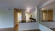 This single-floor residence offers a thoughtfully designed open-concept layout that promotes a natural flow between living spaces. The kitchen serves as a central focal point, featuring crisp white cabinetry that extends to the upper walls, providing ample storage. Dark granite countertops create a striking contrast against the lighter surroundings, while a breakfast bar-style peninsula offers additional workspace and casual seating potential. Stainless steel appliances, including a refrigerator, microwave, and range, complete the functional kitchen setup. A stainless steel sink with a modern faucet is integrated into the peninsula, enhancing both convenience and style. The flooring throughout features warm hardwood-style planks that add continuity and a welcoming feel to the space. Neutral-toned walls are complemented by clean white baseboards and trim, giving the interior a fresh, modern appearance. Track lighting in the kitchen ensures well-lit functionality, while a hallway leading to additional rooms hints at comfortable private living quarters beyond. The overall ambiance is clean, contemporary, and practical, making excellent use of the available space.