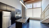 This two-story loft showcases a compelling blend of industrial character and contemporary design. The main level opens into a spacious living area with rich hardwood flooring that reflects natural light streaming in through oversized windows. A sleek, open-concept kitchen anchors the space, featuring dark cabinetry, stainless steel appliances including a range, microwave, and refrigerator, a white subway tile backsplash, and a large central island with a built-in sink and pendant lighting above. The soaring ceilings on this level create an impressive sense of volume, with large floor-to-ceiling windows framing urban views of the surrounding brick buildings. A modern floating staircase with wood treads and a glass railing panel connects the two floors, contributing to the open, airy feel of the main level. The upper level features exposed wood beam ceilings with visible ductwork and piping, lending authentic industrial character to the space. A bedroom on this level is appointed with carpeted flooring, a ceiling fan, and direct access to a private bathroom fitted with dark cabinetry and vanity lighting. Recessed lighting throughout and thoughtful design details complete this distinctive urban loft.