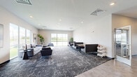 Located at 970 NW 10th St in Waukee, Iowa, this single-floor community amenity facility offers a thoughtfully designed layout spanning a generous footprint. The space is organized into three distinct functional areas, each finished with a consistent modern aesthetic characterized by neutral white walls, dark patterned carpet, and recessed lighting throughout.

The entry hallway features a clean, contemporary reception desk with a light wood countertop and white cabinetry, providing a professional welcome point. Glass-paneled entry doors allow natural light to flood the entrance, creating an open and accessible feel.

The living area serves as the social hub of the facility, offering multiple seating configurations including a lounge zone with plush sofas, accent chairs, and side tables, as well as booth-style seating with tables for more structured gatherings. Large windows frame outdoor views of the surrounding community, further brightening the space. A mounted flat-screen TV and foosball table add to the recreational appeal of the room. Contemporary artwork adorns the walls throughout, adding visual interest.

The exercise room is generously proportioned and well-equipped, housing treadmills, elliptical machines, a cable weight machine, a rowing machine, dumbbells, kettlebells, and a weight bench. Natural light enters through perimeter windows, and overhead fluorescent lighting ensures full visibility across the entire workout floor.