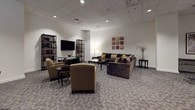 This well-appointed community room is thoughtfully organized into several functional zones, offering residents a versatile shared space for a variety of activities. The lounge area serves as the centerpiece, furnished with a generous dark brown sectional sofa accented by gold and cream throw pillows, complemented by cushioned armchairs and a sleek glass coffee table. Decorative wall art, bookshelf units, and a wall-mounted flat-screen television contribute to the comfortable, residential atmosphere. Recessed lighting illuminates the drop-panel ceiling throughout, casting a warm, even glow across the patterned carpet flooring.

Multiple dining and meeting areas are arranged throughout the room, featuring round tables paired with upholstered chairs, ideal for small group gatherings or collaborative use. Tall indoor plants and framed artwork add natural and visual interest along the walls, enhancing the inviting ambiance.

The fully equipped kitchen is finished with rich mahogany cabinetry, dark granite countertops, and stainless steel appliances including a full-size range and a large French-door refrigerator. Light tile flooring anchors the kitchen area, and a pass-through bar counter with black bar stools creates a seamless connection between the kitchen and the main lounge space.