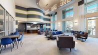 This expansive single-floor amenity space is defined by its dramatic double-height ceilings and bold architectural character. The main lounge area commands attention with sweeping horizontal stripe murals in navy, green, and white wrapping a curved interior wall, complemented by a striking sculptural chandelier overhead. Floor-to-ceiling windows and glass doors along one wall flood the space with natural light while providing views of an outdoor terrace, reinforcing a seamless indoor-outdoor connection.

The lounge is furnished with generous sectional sofas, accent chairs, and small side tables arranged to encourage both casual gatherings and quiet relaxation. A built-in media wall with a large flat-screen television anchors one end of the room, flanked by decorative artwork. Transitioning toward the kitchen area, warm hardwood flooring replaces the neutral carpet, leading to a distinctive curved bar counter finished in rich dark wood with a white surface, barstool seating, and full stainless steel appliances, including a refrigerator and microwave. A dining nook nearby features banquette seating, bold blue accent chairs, and modern pendant lighting, making it ideal for informal dining or socializing.