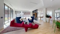 Located at 200 Pleasant St in Malden, MA, this single-floor amenity lounge is a thoughtfully designed communal space that balances modern aesthetics with functional comfort. The open-plan layout is unified by light hardwood flooring and crisp white ceilings with recessed lighting, creating a bright and welcoming atmosphere throughout.

The lounge area anchors the space with bold, contemporary furnishings, including striking crimson sculptural seating, deep navy wingback chairs, and layered area rugs that define distinct relaxation zones. A decorative black-and-white geometric partition elegantly separates the lounge from the adjacent games area, which features a full-size billiards table beneath an artful cluster of geometric pendant lights. Floor-to-ceiling glass walls and doors flood this zone with natural light and provide direct access to an outdoor patio.

The kitchenette area is a standout feature, showcasing warm wood cabinetry complemented by a vivid cobalt blue tile backsplash and a wall-mounted television. A large wood-topped communal counter with vibrant green perforated bar stools provides casual seating. Large-scale potted plants and an abundance of natural light from expansive windows and glass doors leading to a furnished outdoor courtyard complete the inviting, contemporary ambiance.