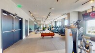 Located along NW Third Ave in Miami's vibrant Wynwood neighborhood, this contemporary fitness center is designed to offer a comprehensive wellness experience across a single, thoughtfully organized floor. The facility is arranged into distinct zones that cater to a variety of exercise disciplines and recovery needs.

The main cardio and strength area features a generous row of treadmills, ellipticals, and stair climbers positioned alongside floor-to-ceiling windows, allowing natural light to flood the space while offering expansive views of the surrounding Miami skyline. A dedicated strength zone houses a full array of cable machines, weight benches, and free weights neatly organized on wall-mounted racks. Large mirrors amplify the sense of space throughout, and track lighting overhead ensures consistent, well-distributed illumination.

A separate enclosed cycling studio accommodates spin bikes arranged facing a mirrored wall, with shelved storage for medicine balls, kettlebells, and exercise accessories. An adjoining studio space features warm hardwood flooring, ideal for yoga, stretching, or group fitness classes.

A welcoming lounge area with wood flooring, blue-green accent walls, curated artwork, and comfortable seating provides a relaxed transition space. Dual water fountains and clearly marked entry points round out the facility's practical amenities, creating a polished and functional environment throughout.