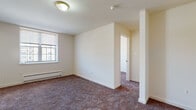 This single-floor residential unit offers a practical and comfortable layout designed to maximize livability. The living area is finished with neutral-toned carpet and crisp white walls, creating a clean and versatile backdrop. A pass-through opening connects the living space to the kitchen, enhancing the sense of openness between the two areas. An electrical panel is conveniently recessed into the living room wall.

The kitchen features warm oak cabinetry extending to the upper and lower sections of the room, complemented by white square tile backsplash, light countertops, and light-toned floor tiles. A gas range and overhead ventilation hood are included, along with a single-basin sink beneath the cabinetry.

The bedrooms are carpeted and share the same neutral color palette seen throughout the unit. Each room is served by baseboard heating and benefits from natural light through window blinds. One bedroom offers a notably bright corner window, while others provide access to an adjoining hallway or secondary room.

The bathroom is finished with full white square wall tile surrounding a soaking bathtub, with a small window positioned above for ventilation and light. The clean, classic tile work gives the bathroom a timeless, functional appearance. Overall, the space presents a straightforward and well-organized layout suited for comfortable everyday living.