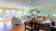 This single-floor space is defined by its open-plan design, where the dining and living areas flow naturally into one another, creating a cohesive and welcoming environment. The warm hardwood flooring runs throughout, grounding the space with a sense of continuity, while soft sage-green walls and a pale blue ceiling lend a calm, coastal-inspired ambiance.

The dining area features a rich, dark-wood table paired with cushioned chairs, offering a comfortable setting for meals and gatherings. Just beyond, the living area is arranged around a large wall-mounted flat-screen television flanked by compact speakers, with a white media console below providing ample storage behind glass-paneled doors. The seating arrangement includes plush accent chairs positioned atop a light, textured area rug, inviting relaxed conversation or quiet leisure.

Crown molding adds a refined architectural detail along the ceiling line, while recessed lighting ensures the interior is evenly and warmly illuminated. Large sliding or French-style glass doors along one wall allow natural light to pour in and offer views of an outdoor seating area, enhancing the sense of openness. Decorative coastal accents are thoughtfully placed throughout, reinforcing the light and airy character of the space.