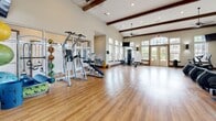 Located at 3333 Hacks Cross Rd in Memphis, Tennessee, this well-appointed fitness center offers a spacious, single-floor layout designed to support a comprehensive workout experience. The facility's most striking architectural feature is its soaring vaulted ceiling adorned with rich, dark-stained exposed wood beams, lending the space a warm yet airy character. Ceiling fans are suspended from the beams, ensuring comfortable airflow throughout.

Natural light floods the interior through an abundance of large windows and arched glass-panel doors, creating a bright and energizing atmosphere. The walls are finished in a clean, neutral tone, complemented by full-length mirrors that further enhance the sense of openness. Durable hardwood-style flooring runs throughout the main exercise area, providing both aesthetics and functionality.

The cardio section is well-equipped with multiple treadmills and elliptical trainers, each stationed on protective floor mats. The strength training area features a cable machine, a dedicated bench press station, and a medicine ball rack stocked with a variety of weighted balls. A wall-mounted flat-screen TV serves the cardio zone, and additional screens are positioned throughout the space. A water fountain and sanitation station are conveniently available, rounding out this fully functional and thoughtfully designed fitness facility.