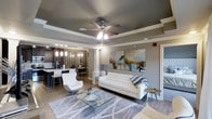 This single-floor space showcases a contemporary open-concept design that connects the living and dining areas in a cohesive, flowing layout. The living area is anchored by a tray ceiling finished in a deep charcoal gray, adding architectural depth and contrast against the neutral gray walls. A ceiling fan with a built-in light fixture serves as a functional centerpiece overhead. Decorative columns frame the transition between spaces, adding a structural elegance to the floor plan. Large-scale abstract artwork in vivid blues and yellows provides a striking focal point on one wall, while the hardwood-style flooring runs throughout, unifying the space. The living area is furnished with a crisp white tufted sofa, a modern swivel accent chair, and a glass-top coffee table, all arranged atop a patterned area rug. Adjacent to the living space, the dining area features pendant lighting and a decorative mirrored wall accent, creating visual intrigue. The kitchen beyond is equipped with dark cabinetry and a light countertop with bar seating. Natural light filters in through windows, brightening the overall ambiance and enhancing the modern, polished aesthetic throughout.