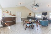 Located at 200 Red Cedar Ct in Chesapeake, Virginia, this single-story community clubhouse offers a thoughtfully designed gathering space that balances comfort and functionality. The open-plan interior is anchored by soaring vaulted ceilings that lend an airy, expansive feel throughout.

The main lounge area is a standout, featuring a mosaic-tiled fireplace flanked by large windows that flood the room with natural light. Wall-mounted television, vibrant area rugs, and a mix of mid-century modern furnishings — including a charcoal sofa, teal accent chairs, and a wooden sideboard topped with a round decorative mirror — create a warm and inviting atmosphere.

Adjacent to the lounge is a casual dining area centered around a round glass-top table with mid-century wooden chairs. Sliding glass doors and large windows on multiple walls provide generous views of the surrounding outdoor greenery and easy access to the exterior.

A separate office or business center is accessible via glass-paned French doors, offering a more private and functional workspace. Track lighting throughout the space ensures consistent, well-distributed illumination. Light tile flooring runs seamlessly across the entire layout, tying the rooms together cohesively.

Overall, this clubhouse presents a bright, welcoming environment well-suited for community gatherings, casual meetings, and everyday resident use.