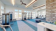 Located in New Port Richey, FL, this fitness center offers a generously sized, single-floor exercise space designed to support a comprehensive workout experience. The room features soaring ceilings with an elegant tray detail and modern linear pendant lighting, complemented by multiple ceiling fans that maintain a comfortable environment during workouts. Large double-pane windows line the walls in stacked configurations, flooding the interior with natural light and offering pleasant views of the surrounding neighborhood. The white shiplap wall paneling adds a clean, contemporary coastal aesthetic throughout.

The flooring features a distinctive two-tone carpet tile design in teal and light grey geometric patterns, visually delineating different workout zones. A full wall of mirrors enhances the sense of space and supports proper form during exercise. The facility is outfitted with a diverse array of professional-grade equipment, including power racks, cable machines, Matrix benches, treadmills, stair climbers, stationary bikes, and a comprehensive free weight section with kettlebells, medicine balls, and dumbbells. A plyo box and stretching area round out the functional training options. A small reception counter near the entrance and a glass-panel door provide a welcoming, organized entry point to the space.