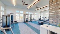 Located in New Port Richey, FL, this fitness center offers a generously sized, single-floor exercise space designed to support a comprehensive workout experience. The room features soaring ceilings with an elegant tray detail and modern linear pendant lighting, complemented by multiple ceiling fans that maintain a comfortable environment during workouts. Large double-pane windows line the walls in stacked configurations, flooding the interior with natural light and offering pleasant views of the surrounding neighborhood. The white shiplap wall paneling adds a clean, contemporary coastal aesthetic throughout.

The flooring features a distinctive two-tone carpet tile design in teal and light grey geometric patterns, visually delineating different workout zones. A full wall of mirrors enhances the sense of space and supports proper form during exercise. The facility is outfitted with a diverse array of professional-grade equipment, including power racks, cable machines, Matrix benches, treadmills, stair climbers, stationary bikes, and a comprehensive free weight section with kettlebells, medicine balls, and dumbbells. A plyo box and stretching area round out the functional training options. A small reception counter near the entrance and a glass-panel door provide a welcoming, organized entry point to the space.